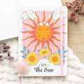 Notebook with a sun design and 'The Sun' title on a soft surface with flowers.