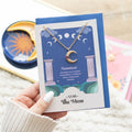 Necklace with a moon charm on a card held by a hand, with a blurred background