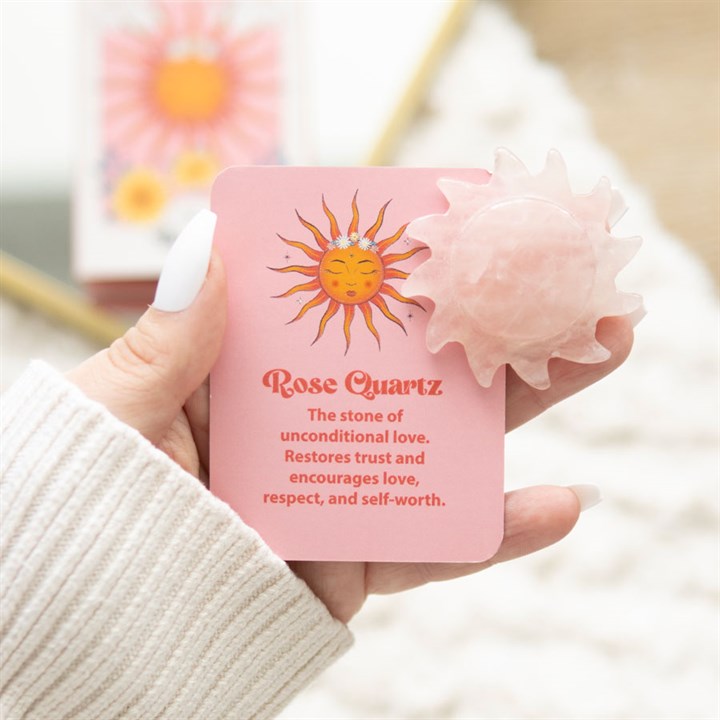 Hand holding a pink card with a sun illustration and text about Rose Quartz, against a blurred background.