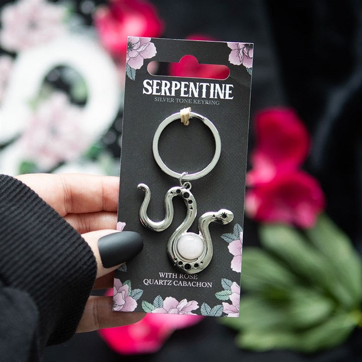 Serpentine keychain packaging held by a hand with black nail polish, against a blurred floral background.