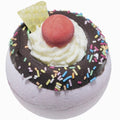 Bath bomb with chocolate topping, whipped cream, and colorful sprinkles on a white background