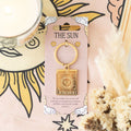 Gold keychain with 'The Sun' design on a pink card, surrounded by decorative elements.
