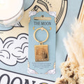 Gold keychain with 'The Moon' card design on a blue card, with decorative elements in the background.
