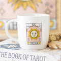 White mug with 'The Sun' tarot card design on a surface with 'The Book of Tarot' visible.