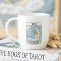 White mug with 'The Moon' tarot card design on a book titled 'The Book of Tarot'.