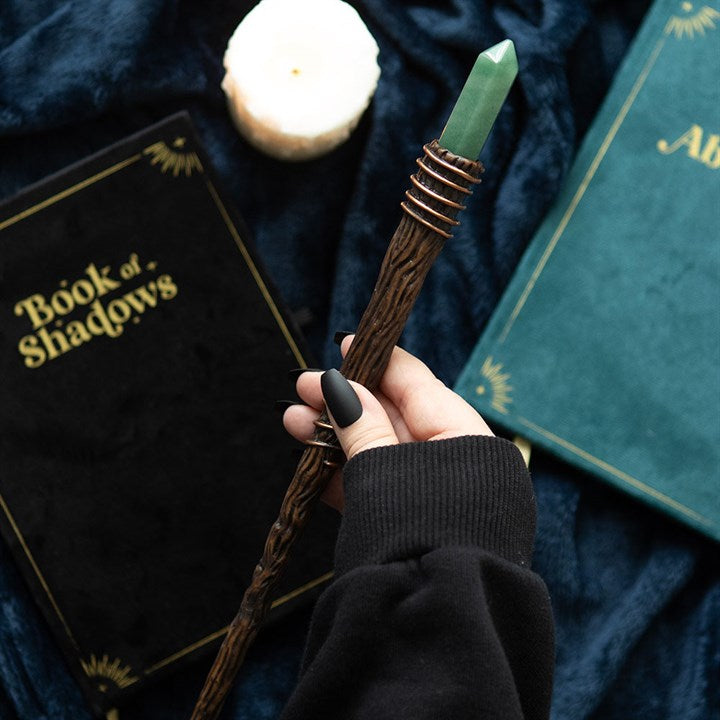 Hand holding a wand with books titled 'Book of Shadows' and 'Alb' in the background.