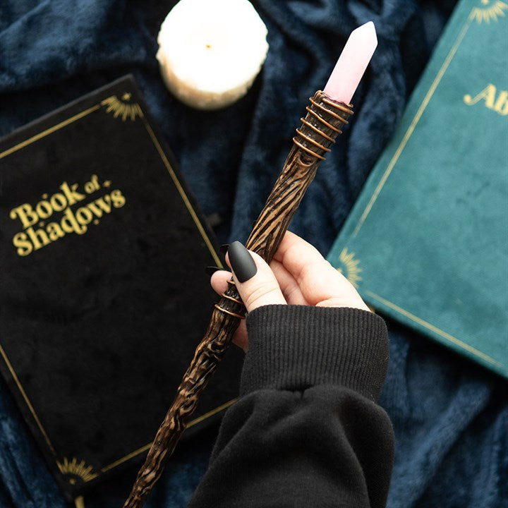 Hand holding a wooden wand with a pink top, surrounded by books titled 'Book of Shadows' and 'Alb'.
