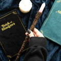 Hand holding a wooden wand with a pink top, surrounded by books titled 'Book of Shadows' and 'Alb'.