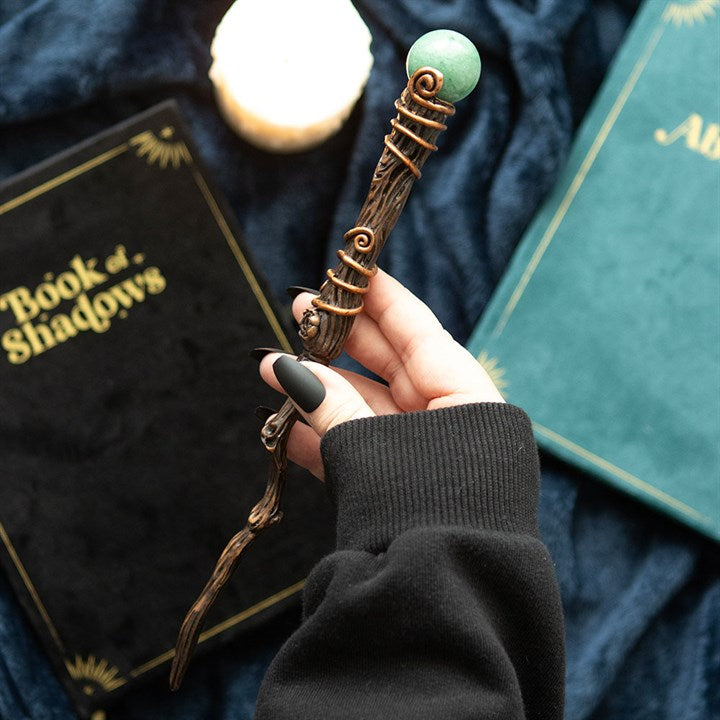 Hand holding a decorative wand with a green gem, against a dark background with books.